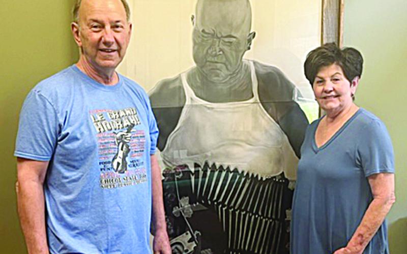 Gilbert “Winky” Aucoin (left) and his wife, Margaret “Chicken,” are pictured alongside an artist’s rendition of the late Cajun music performer Cyprien Landreneau, who is the father of Mrs. Aucoin. (Gazette photo by Tony Marks)